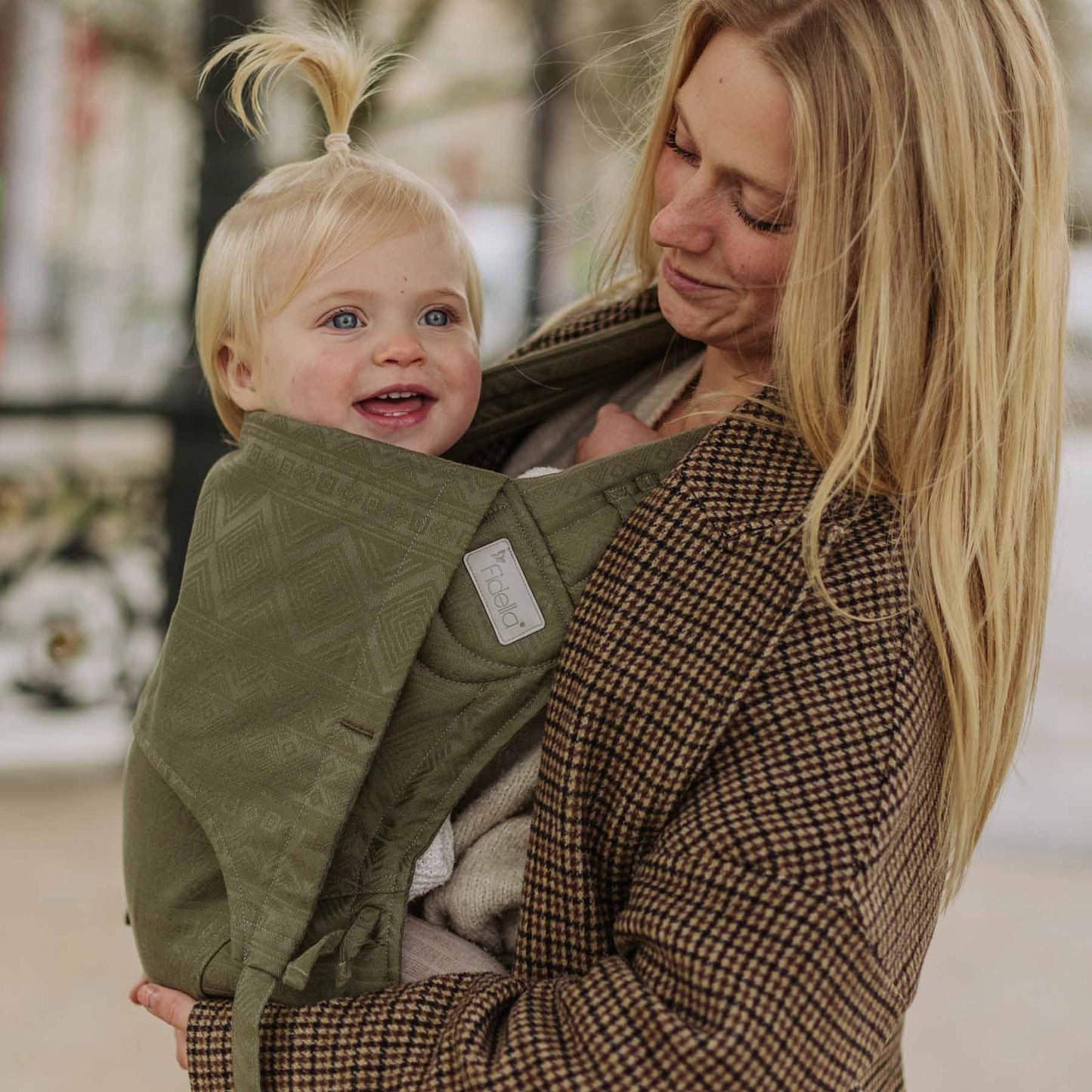 A baby is carried in Fly Tai Mei Tai Sling baby carrier Design Cubic Lines moss