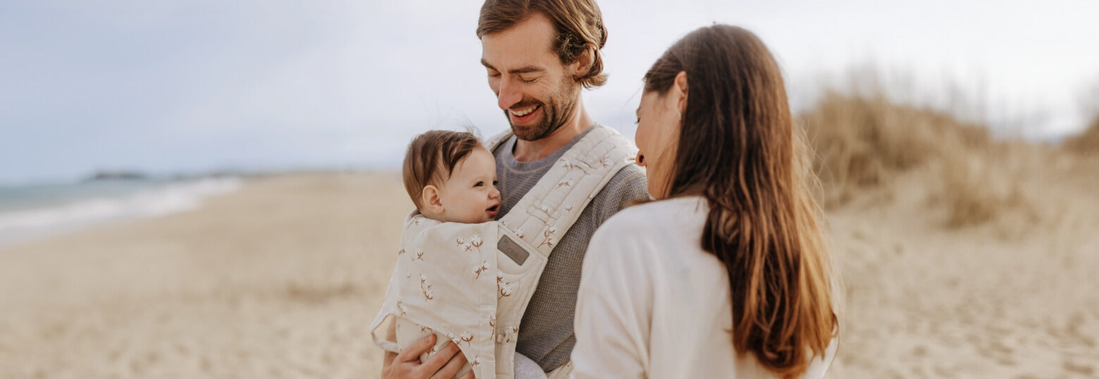 A father is carrying his baby in a FlyTai cotton flower baby carrier at the beach. The mother and the baby are smiling at each other