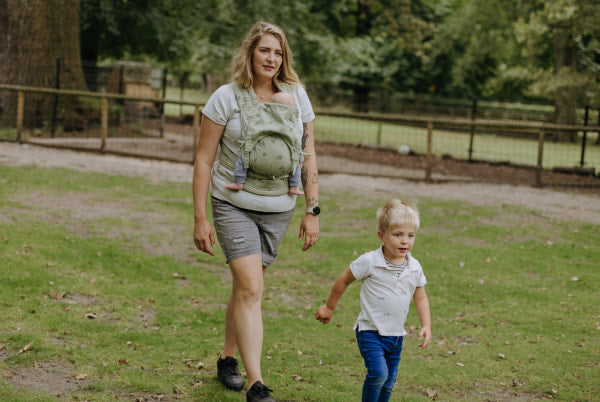 A mother is carrying her baby in a FlyClick clover baby carrier in a park. A toddler is leading the way
